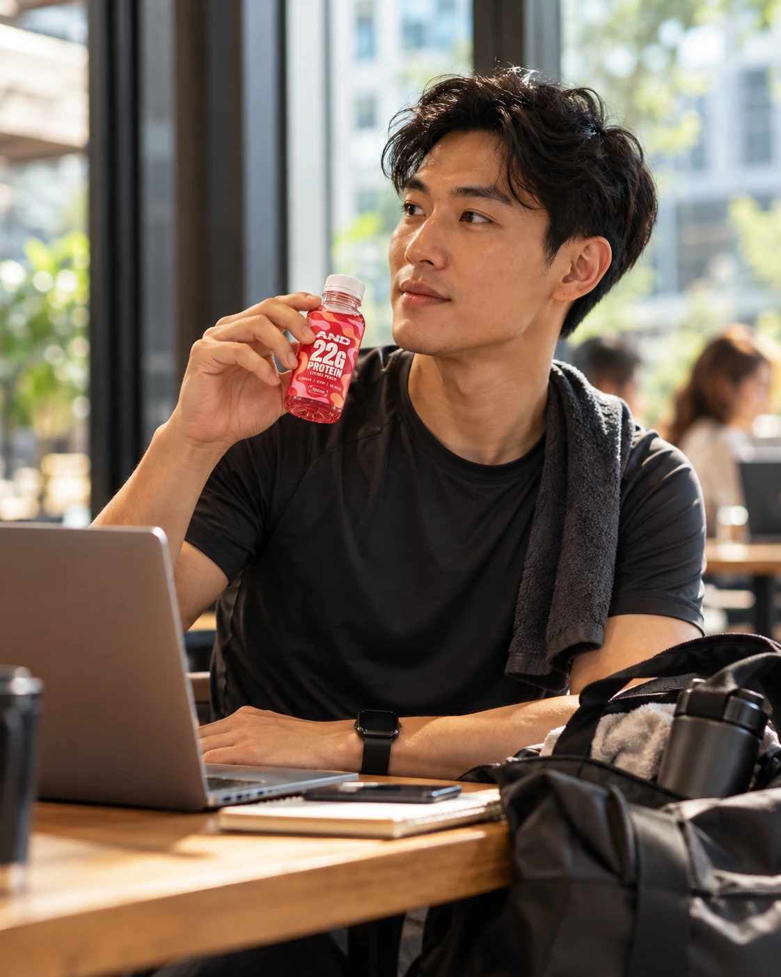 A man in gym clothes at a laptop by a window, holding an AND+ Protein Shot